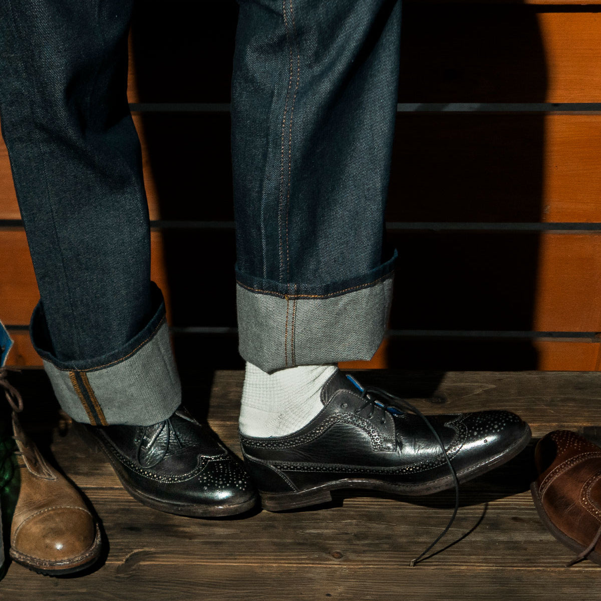 A person in rolled-up jeans and white socks wears the Bed|Stü Sandro II, a black handcrafted leather dress shoe with one shoelace untied. Unique wingtip broguing details add a touch of elegance to its old-world construction. Other shoes can be seen on the wooden floor nearby.