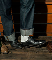 A person in rolled-up jeans and white socks wears the Bed|Stü Sandro II, a black handcrafted leather dress shoe with one shoelace untied. Unique wingtip broguing details add a touch of elegance to its old-world construction. Other shoes can be seen on the wooden floor nearby.