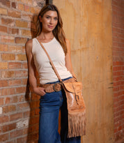 A woman in a white sleeveless top and blue jeans leans against a brick wall, wearing the Bed|Stü Sandy Lane II, a handmade tan fringe crossbody bag crafted from vegetable-tanned leather, along with a brown belt.