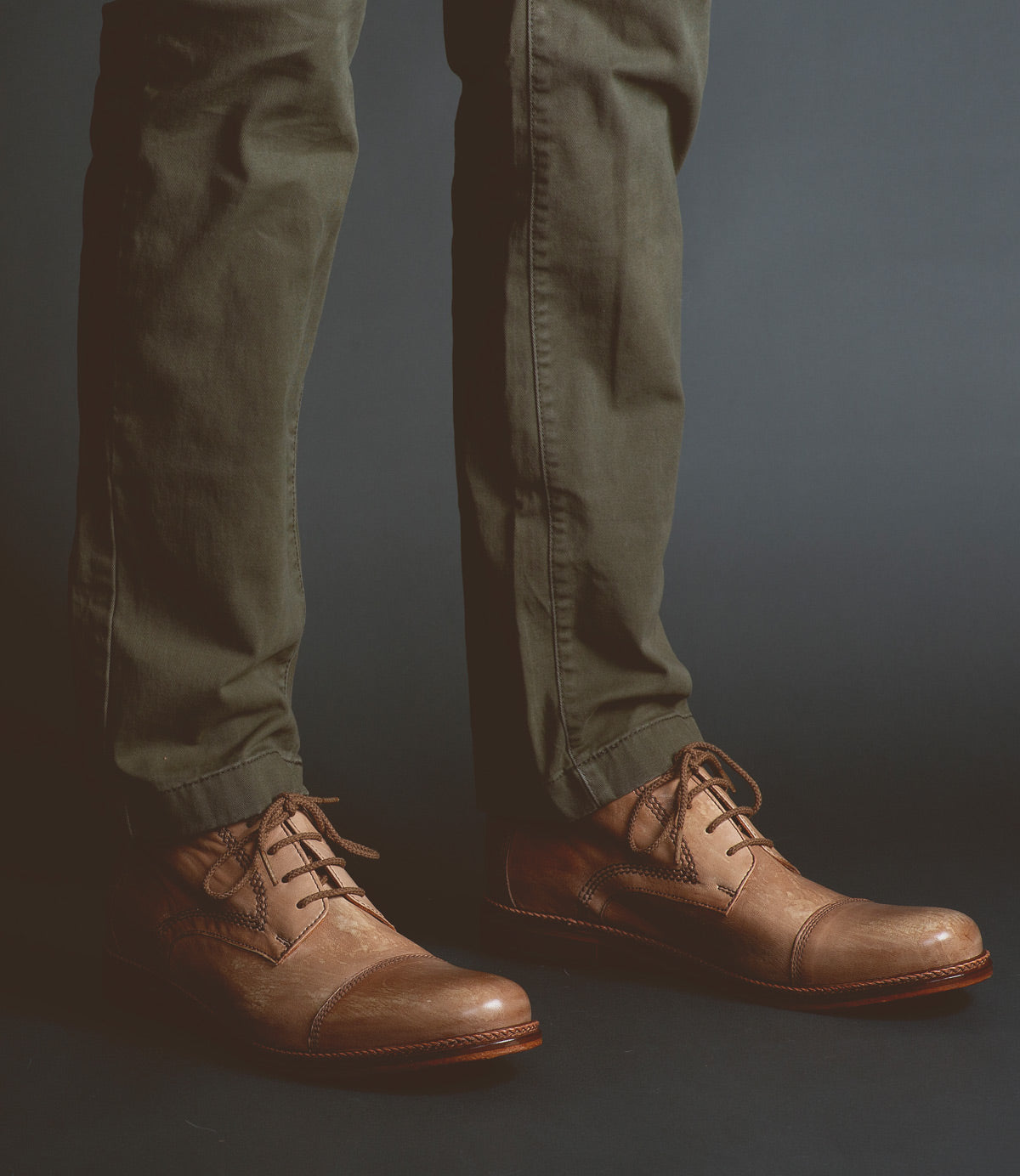 A person wearing olive green pants and the Scoot brown leather Oxford shoes by Bed|Stü, handcrafted in Italy from quality leather, stands on a dark background.