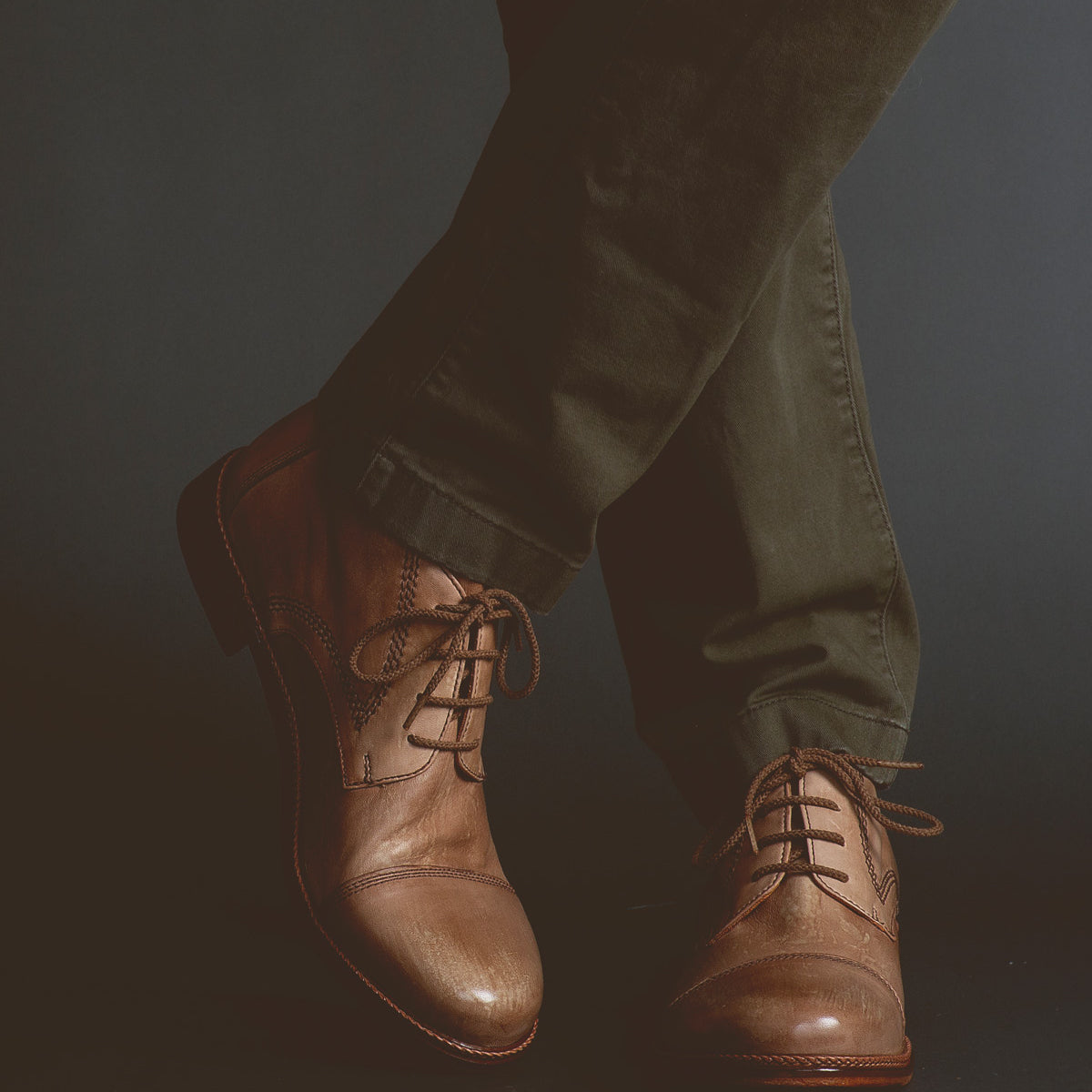 A person wearing olive green pants and the Scoot brown leather lace-up boots by Bed|Stü, handcrafted in Italy, stands with one leg crossed over the other against a dark background.