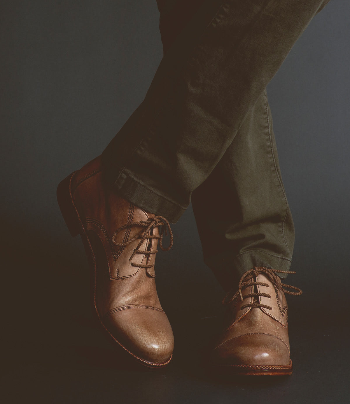 A person wearing olive green pants and the Scoot brown leather lace-up boots by Bed|Stü, handcrafted in Italy, stands with one leg crossed over the other against a dark background.
