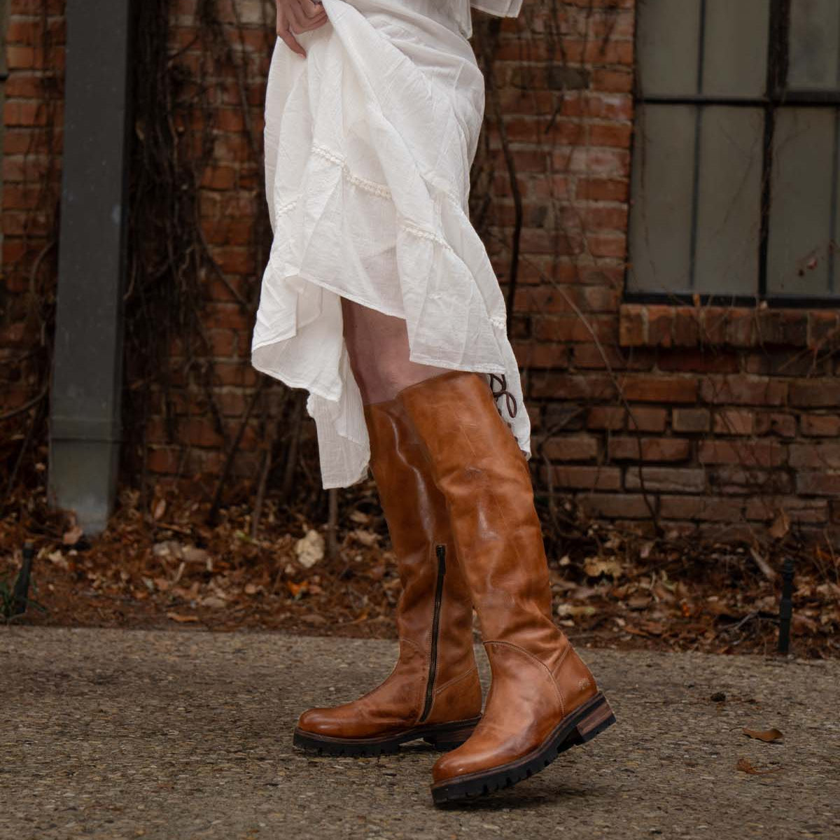 A person wearing the Scope tall vegetable-tanned leather boots by Bed|Stü and a white dress stands on a stone path in front of a brick wall and window.