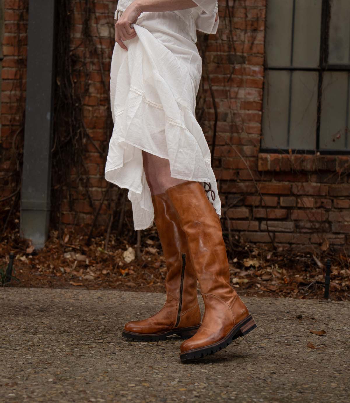 A person wearing the Scope tall vegetable-tanned leather boots by Bed|Stü and a white dress stands on a stone path in front of a brick wall and window.