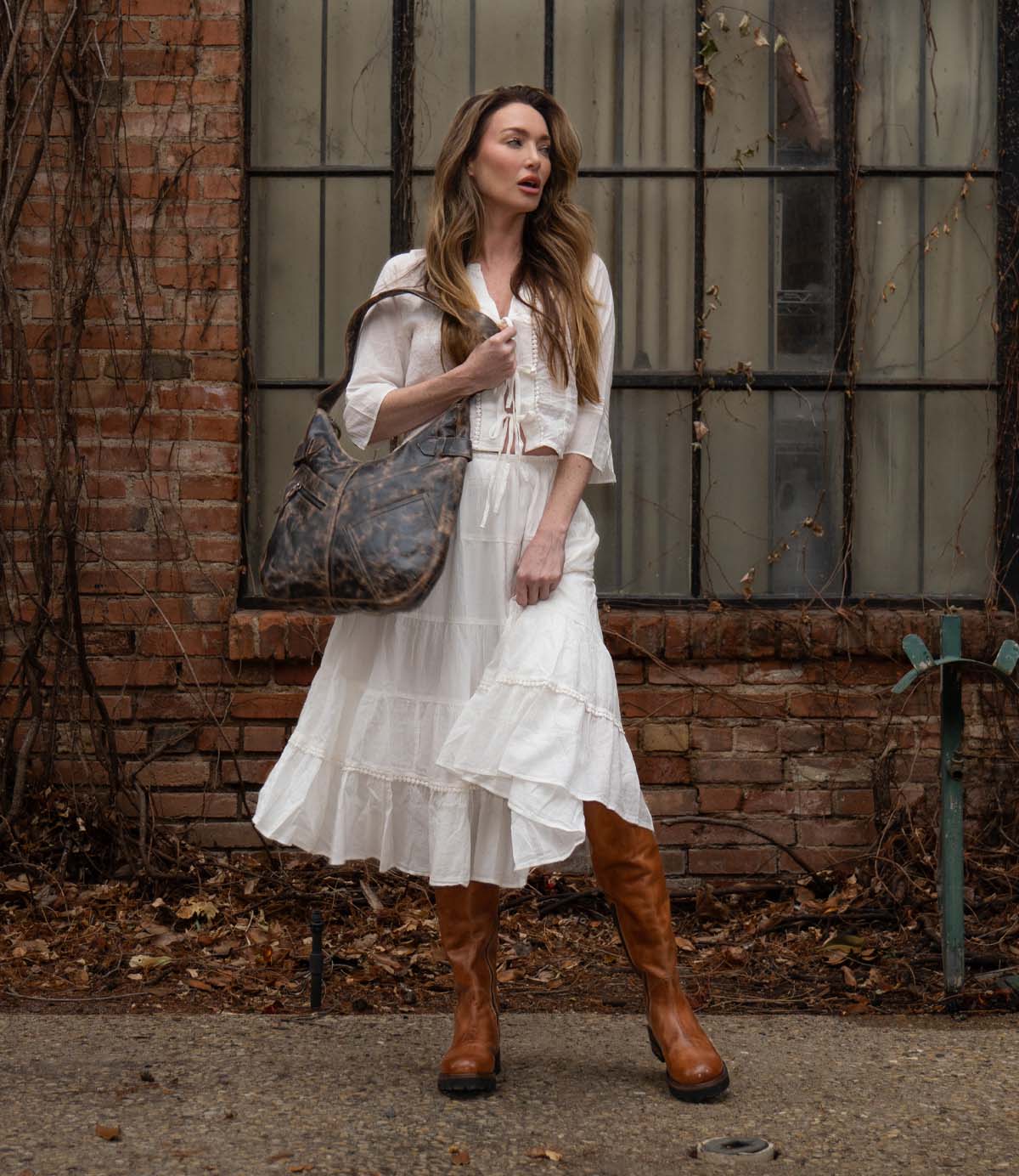 A woman in a white dress stands in front of a brick wall with large windows, holding a large brown bag. Her Scope tall leather boots by Bed|Stü, expertly crafted from vegetable-tanned leather, add a timeless touch to her elegant ensemble.