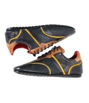 A pair of Bed|Stü Scurry black retro-inspired sneakers, crafted from vegetable-tanned leather with yellow and brown accents, red inner lining, and textured rubber soles for everyday durability, are displayed side by side.