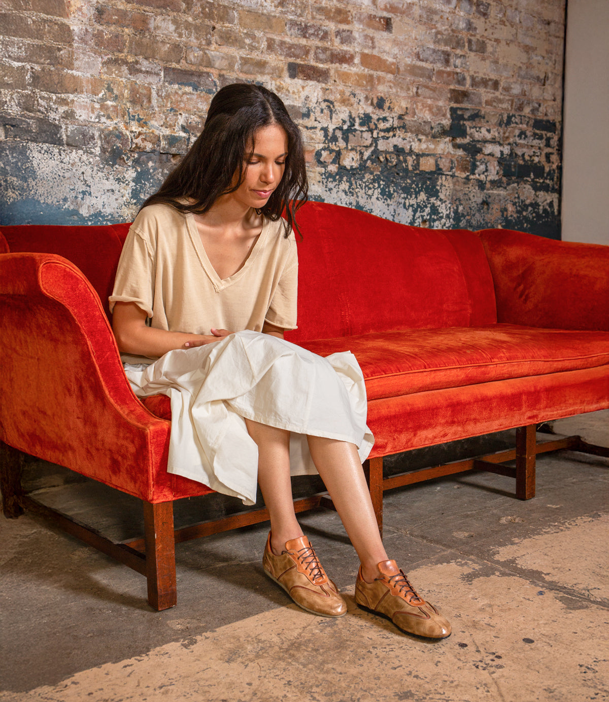A woman sits on a red velvet couch against a worn brick wall, looking down with her hands in her lap. She wears a beige top, white skirt, and Bed|Stü's Scurry sneakers—tan retro-inspired shoes crafted from vegetable-tanned leather that add an understated touch of everyday durability to her look.