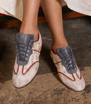 Someone is wearing Bed|Stü Scurry II lace-up shoes in beige and gray with red accents, sitting indoors on a concrete floor.