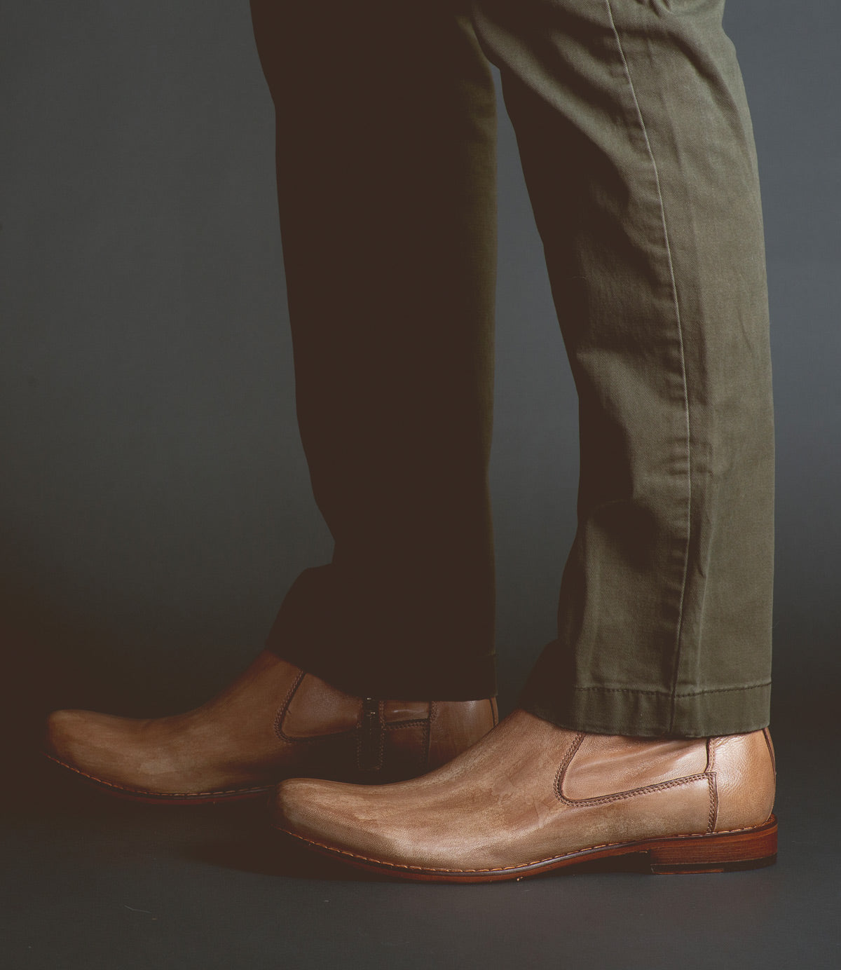 A person wearing olive green pants and light brown Seek Chelsea boots by Bed|Stü, handmade in Italy from vegetable-tanned leather, stands on a dark surface.