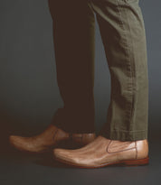 A person wearing olive green pants and light brown Seek Chelsea boots by Bed|Stü, handmade in Italy from vegetable-tanned leather, stands on a dark surface.