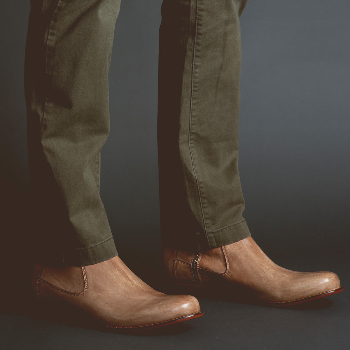 Wearing the Bed|Stü Seek brown vegetable-tanned leather slip-on shoes and olive green pants, a person stands against a dark background.