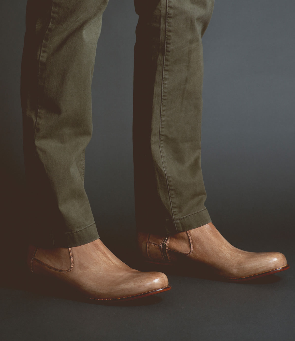 Wearing the Bed|Stü Seek brown vegetable-tanned leather slip-on shoes and olive green pants, a person stands against a dark background.