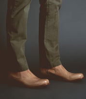 Wearing the Bed|Stü Seek brown vegetable-tanned leather slip-on shoes and olive green pants, a person stands against a dark background.