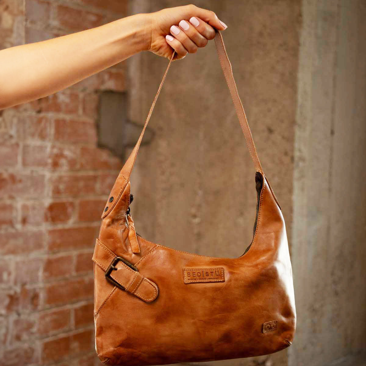 A hand holds the Seemly by Bed|Stü, a brown leather shoulder bag featuring a buckle detail and embossed branding; its compact shape makes it ideal for everyday styling, set against a brick and concrete background.