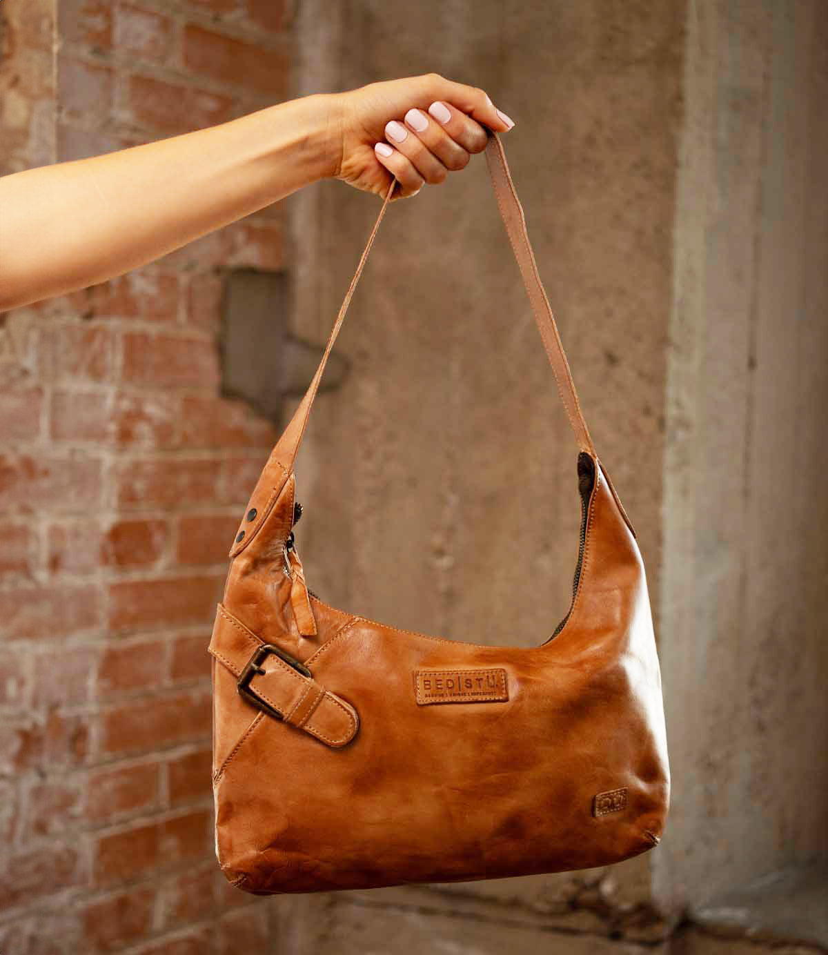 A hand holds the Seemly by Bed|Stü, a brown leather shoulder bag featuring a buckle detail and embossed branding; its compact shape makes it ideal for everyday styling, set against a brick and concrete background.