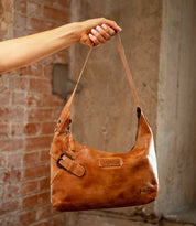 A hand holds the Seemly by Bed|Stü, a brown leather shoulder bag featuring a buckle detail and embossed branding; its compact shape makes it ideal for everyday styling, set against a brick and concrete background.