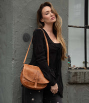 A woman with long hair, dressed in a black blouse and patterned pants, stands against a gray wall. She carries the Bed|Stü Sequoia EM, a brown vegetable-tanned leather saddle bag, over her shoulder and gazes slightly past the camera.