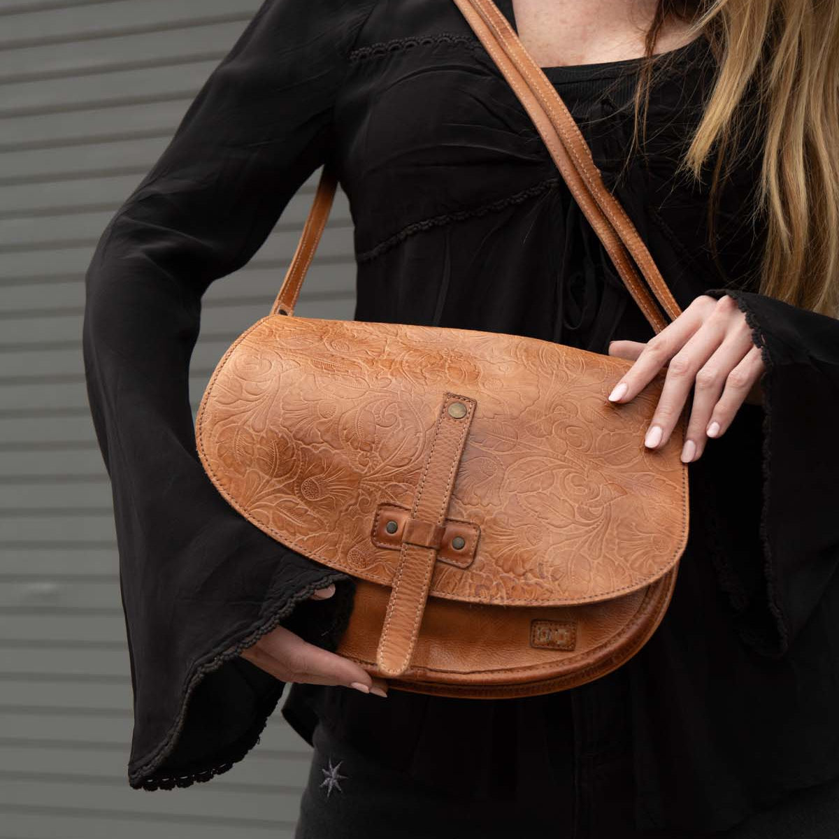 A person with long hair wears a black top and holds the Bed|Stü Sequoia EM vegetable-tanned leather shoulder bag in tan against a gray, striped background.