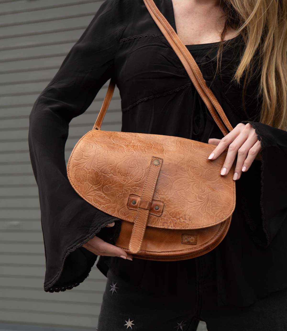 A person with long hair wears a black top and holds the Bed|Stü Sequoia EM vegetable-tanned leather shoulder bag in tan against a gray, striped background.