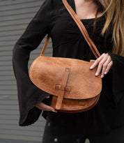 A person with long hair wears a black top and holds the Bed|Stü Sequoia EM vegetable-tanned leather shoulder bag in tan against a gray, striped background.