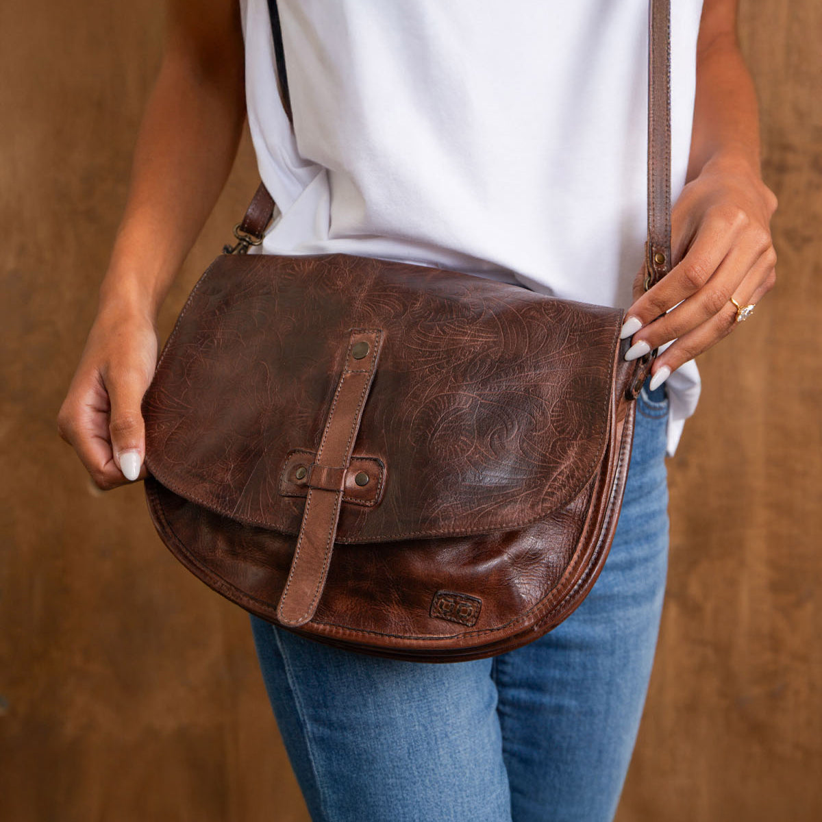 A person wearing a white top and blue jeans holds the Bed|Stü Sequoia EM, a brown vegetable-tanned leather saddle bag with a buckle detail, in front of a wooden background.