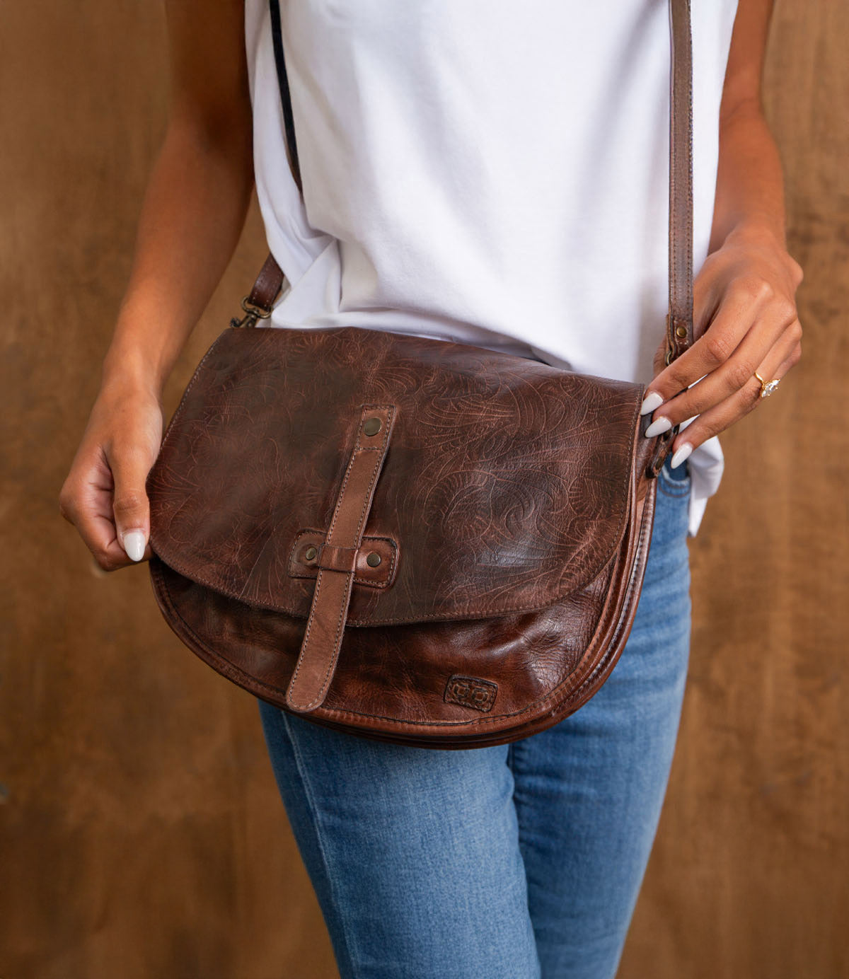 A person wearing a white top and blue jeans holds the Bed|Stü Sequoia EM, a brown vegetable-tanned leather saddle bag with a buckle detail, in front of a wooden background.