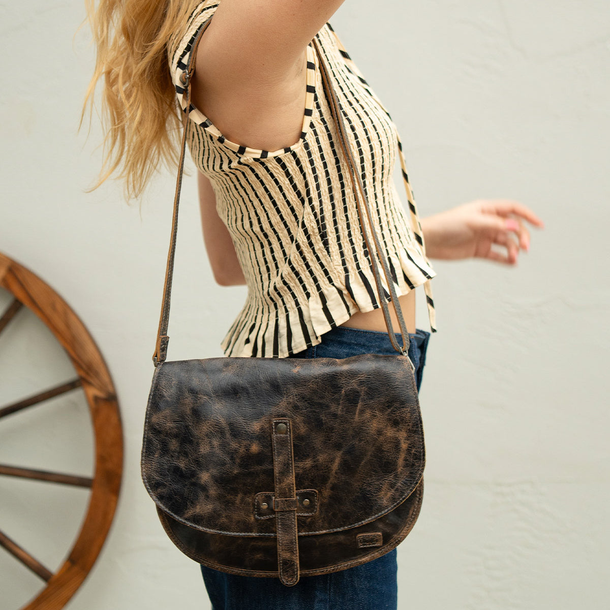 A person wearing a sleeveless striped top and jeans holds their arm up, carrying the Bed|Stü Sequoia—a worn brown vegetable-tanned leather crossbody bag with a sturdy strap—while a wooden wheel sits in the background.