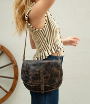 A person wearing a sleeveless striped top and jeans holds their arm up, carrying the Bed|Stü Sequoia—a worn brown vegetable-tanned leather crossbody bag with a sturdy strap—while a wooden wheel sits in the background.