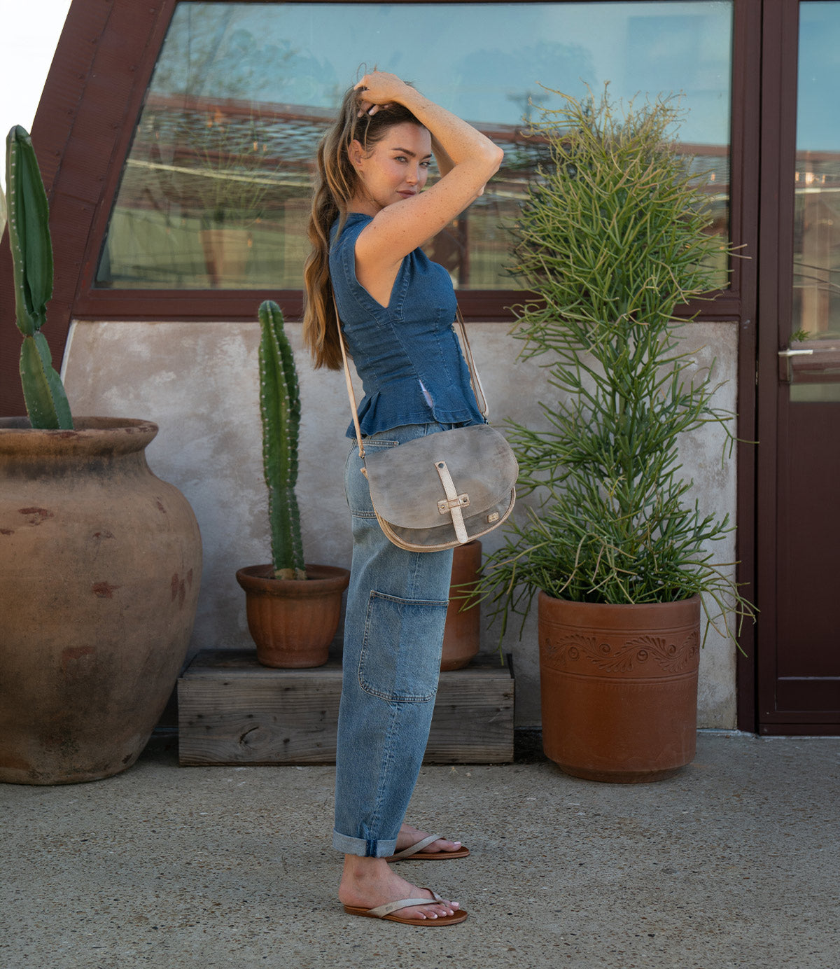 A woman stands outdoors near potted plants, wearing a blue denim top, jeans, and sandals. Slung effortlessly on her shoulder is the Bed|Stü Sequoia—a gray vegetable-tanned leather bag featuring a sleek tab closure and a comfortable crossbody strap. As she adjusts her hair with one hand, the Sequoia complements her relaxed yet stylish look.