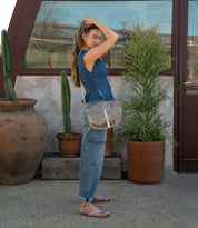 A woman stands outdoors near potted plants, wearing a blue denim top, jeans, and sandals. Slung effortlessly on her shoulder is the Bed|Stü Sequoia—a gray vegetable-tanned leather bag featuring a sleek tab closure and a comfortable crossbody strap. As she adjusts her hair with one hand, the Sequoia complements her relaxed yet stylish look.