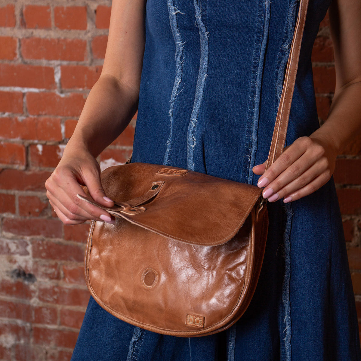 A person in a blue denim dress holds the Bed|Stü Sequoia, a brown leather crossbody bag with a crossbody strap and tab closure, in front of a red brick wall.