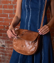 A person in a blue denim dress holds the Bed|Stü Sequoia, a brown leather crossbody bag with a crossbody strap and tab closure, in front of a red brick wall.
