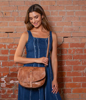 A woman in a sleeveless blue dress stands against a brick wall, holding the Bed|Stü Sequoia vegetable-tanned leather crossbody bag with one hand, its tab closure visible, and smiling slightly.