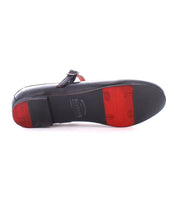 The Bed|Stü Shuffle III is a black leather dress shoe showcased from the sole, highlighting its distinctive red and black rubber tread sections, branded detailing, and a strap accented with a metal buckle. Crafted from supple leather for enduring comfort, this style artfully merges classic masculine lines with the refined detailing typically associated with feminine footwear.