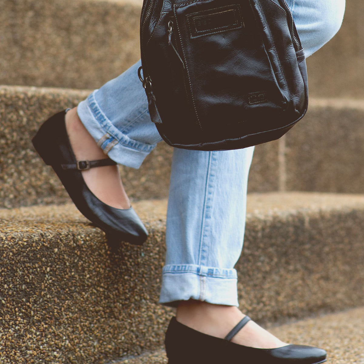 A person wearing light blue jeans and black Mary Jane shoes—an elegant take on feminine footwear—sits on concrete steps, holding the Shuffle III supple leather black backpack by Bed|Stü.