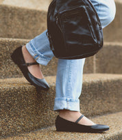 A person wearing light blue jeans and black Mary Jane shoes—an elegant take on feminine footwear—sits on concrete steps, holding the Shuffle III supple leather black backpack by Bed|Stü.