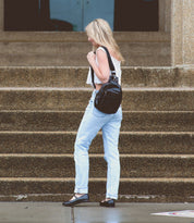 A woman stands on a wet sidewalk in front of stone steps, wearing light blue jeans, a white top, and black Bed|Stü Shuffle III ballet flats, and carrying a large black shoulder bag.