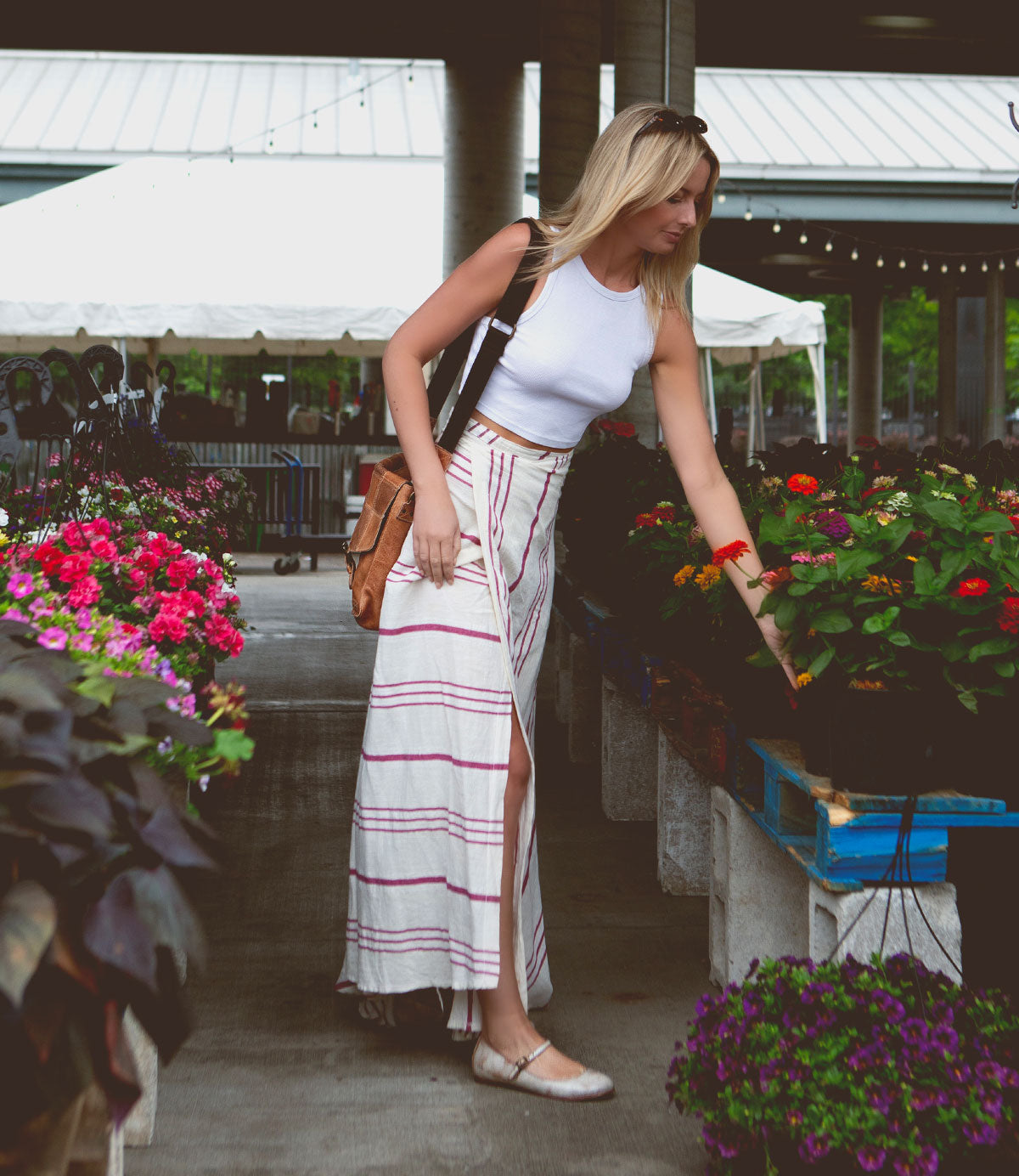 A woman in a white tank top and striped skirt, wearing the supple leather Shuffle III ballet flats by Bed|Stü, examines potted flowers at an outdoor market, surrounded by colorful blooms and plants.
