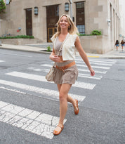 A woman in a beige outfit and Bed|Stü Shuffle III supple leather ballet flats walks across a city crosswalk, smiling. Tall buildings and a few pedestrians are in the background.