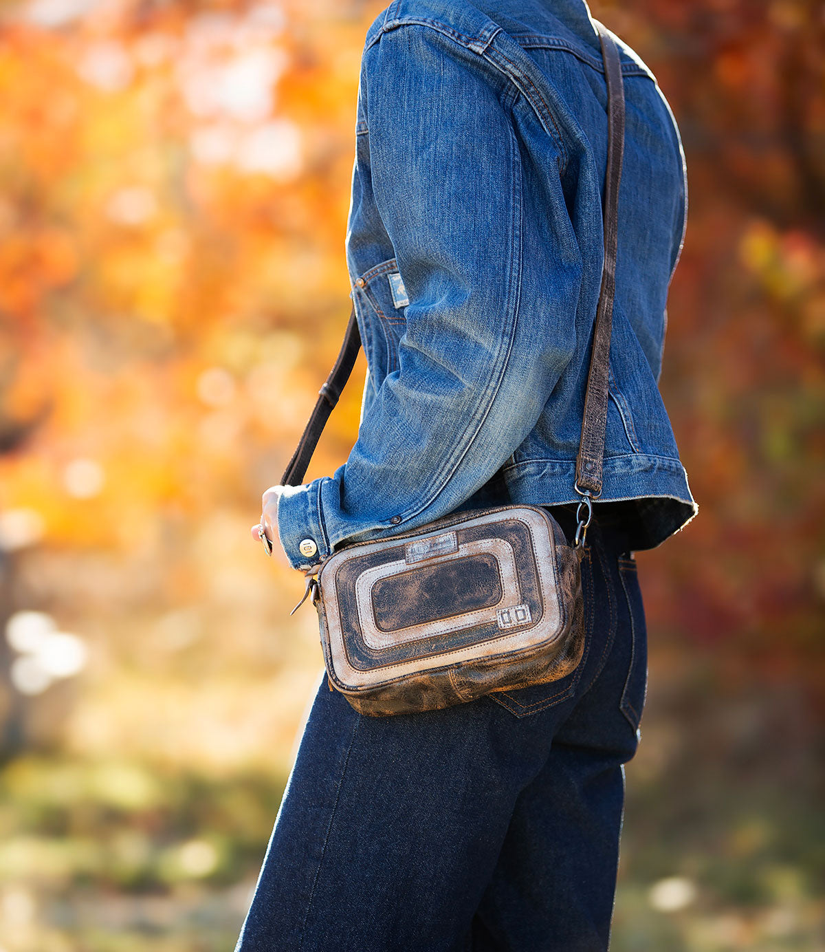 A person stands outdoors against blurred autumn foliage, wearing a blue denim jacket and jeans, and carrying the Bed|Stü Shy purse—an organized bag featuring dual pockets.