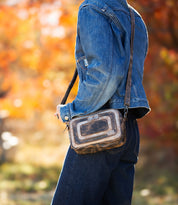 A person stands outdoors against blurred autumn foliage, wearing a blue denim jacket and jeans, and carrying the Bed|Stü Shy purse—an organized bag featuring dual pockets.