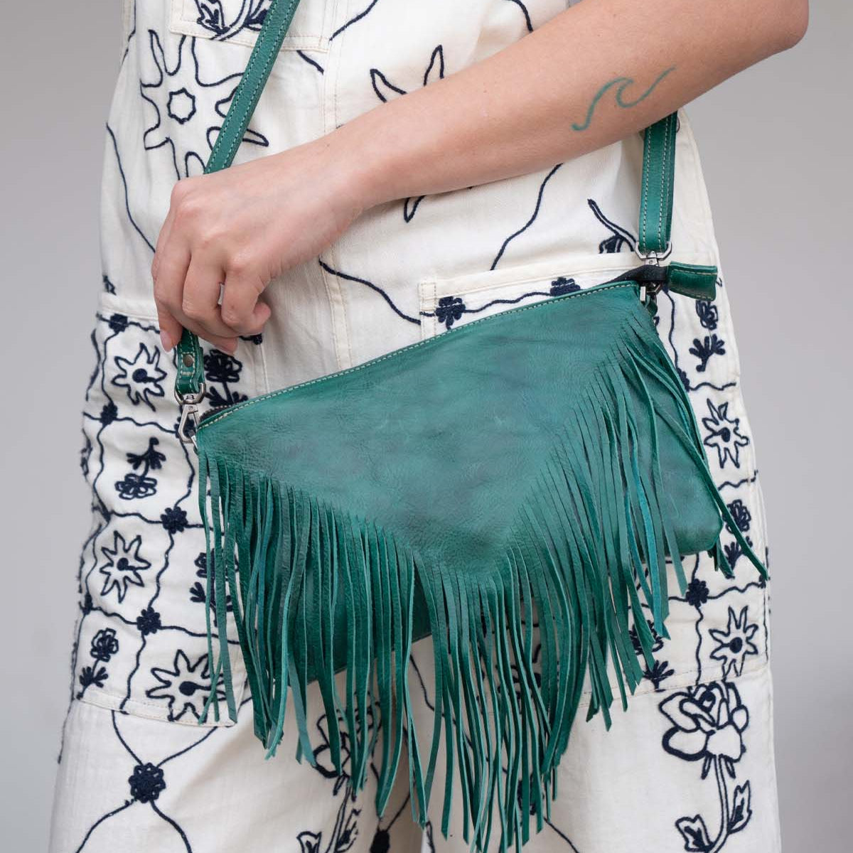 A person dressed in a white outfit with blue floral patterns is holding the Bed|Stü Silverrock green fringe flap bag, with a wave tattoo visible on their forearm.