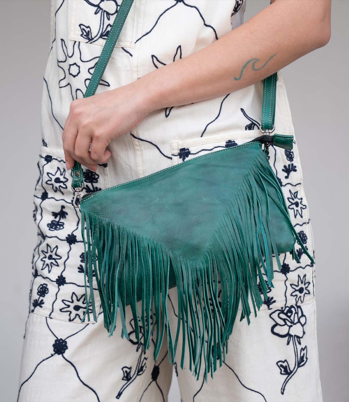 A person dressed in a white outfit with blue floral patterns is holding the Bed|Stü Silverrock green fringe flap bag, with a wave tattoo visible on their forearm.