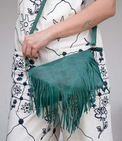 A person dressed in a white outfit with blue floral patterns is holding the Bed|Stü Silverrock green fringe flap bag, with a wave tattoo visible on their forearm.