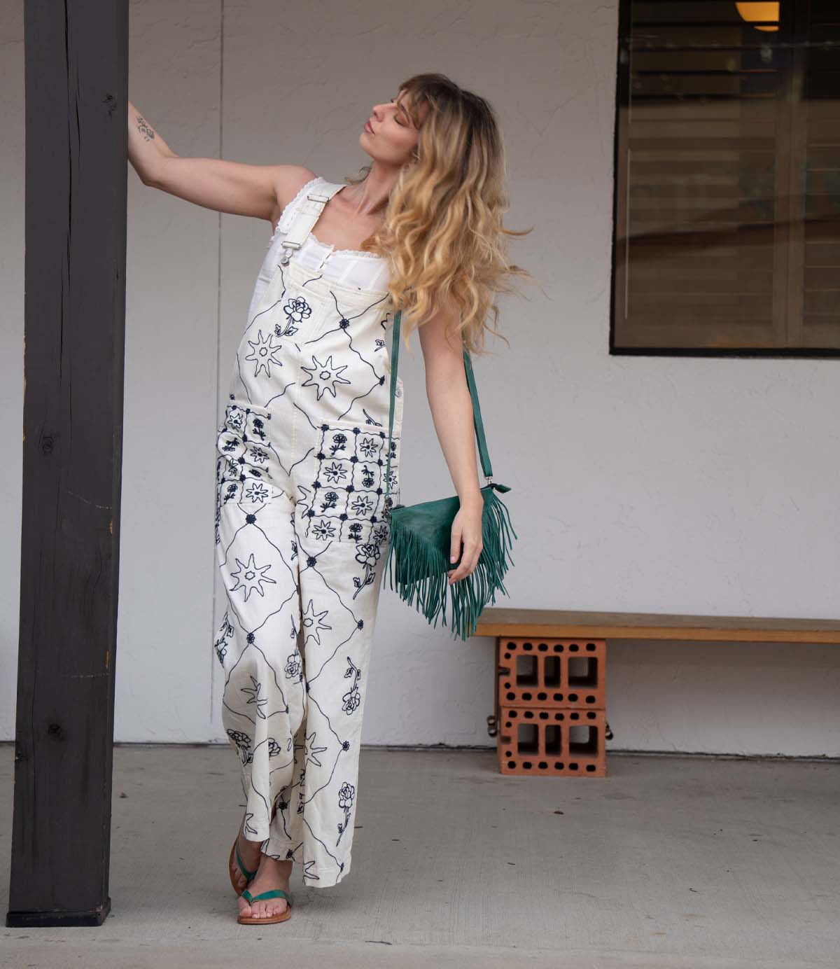 A woman in patterned white overalls and sandals stands by a dark post, holding it with one hand. She carries the Bed|Stü Silverrock fringe flap bag—a chic green accessory—and poses in front of a white wall and wooden bench.