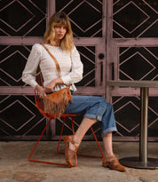 A woman in a white eyelet top, blue jeans, and tan leather shoes sits on an orange chair, holding the Bed|Stü Silverrock small crossbody bag featuring a fringe flap detail.