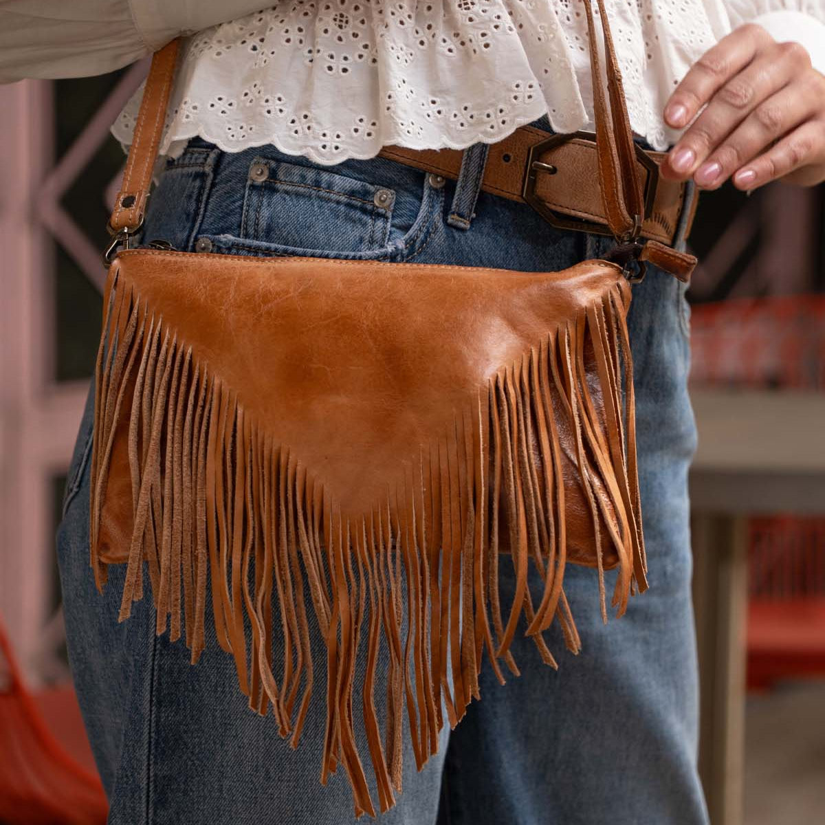 A person wearing blue jeans, a white eyelet blouse, and a brown belt holds the Bed|Stü Silverrock—a stylish small crossbody bag crafted in brown leather with fringe and a flap design.