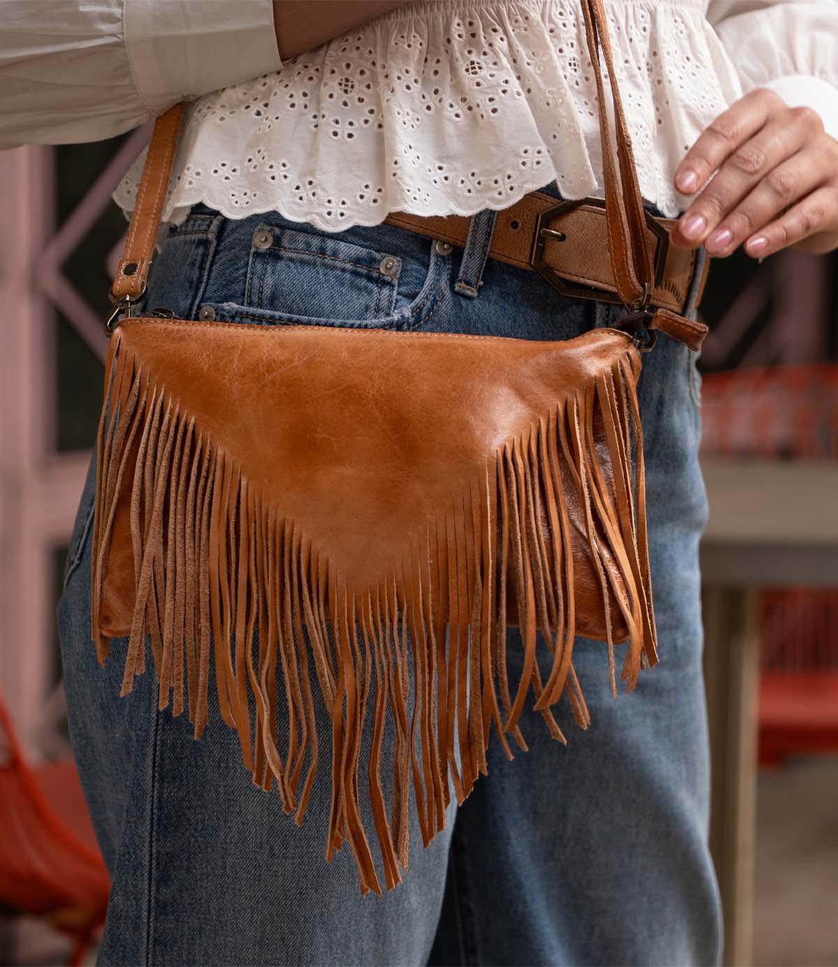 A person wearing blue jeans, a white eyelet blouse, and a brown belt holds the Bed|Stü Silverrock—a stylish small crossbody bag crafted in brown leather with fringe and a flap design.