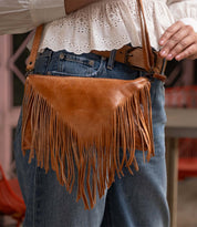 A person wearing blue jeans, a white eyelet blouse, and a brown belt holds the Bed|Stü Silverrock—a stylish small crossbody bag crafted in brown leather with fringe and a flap design.