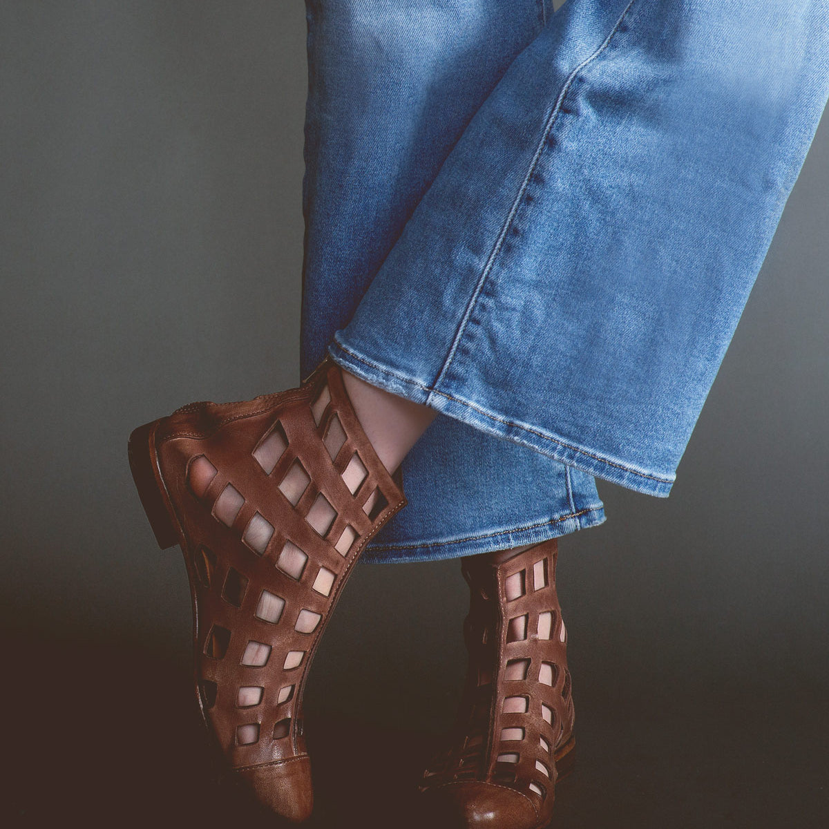 Wearing the Bed|Stü Skyline brown ankle boots handcrafted in Italy, a person in blue jeans stands with legs crossed against a dark background.